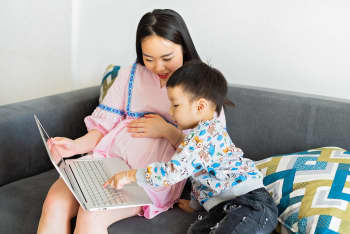 A pregnant mother sits with her young son on a couch as they use a laptop together, enjoying quality time indoors.