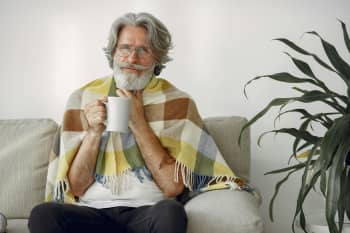 Elderly man wrapped in a shawl sitting on a couch holding a mug, indoors.